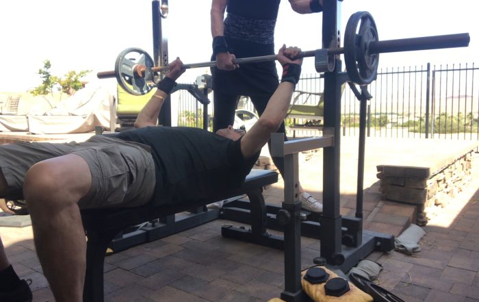 Personal strength training with barbell bench press and hands-on strength coaching for an older adult.